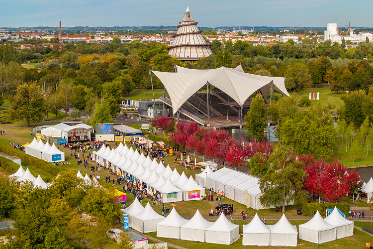 Luftaufnahme des Explore Science Geländes im Elbauenpark Magdeburg mit weißen Pagodenzelten, Bühnenbereich und Besucher:innen vor der markanten Seebühne und dem Jahrtausendturm, umgeben von herbstlich gefärbten Bäumen.